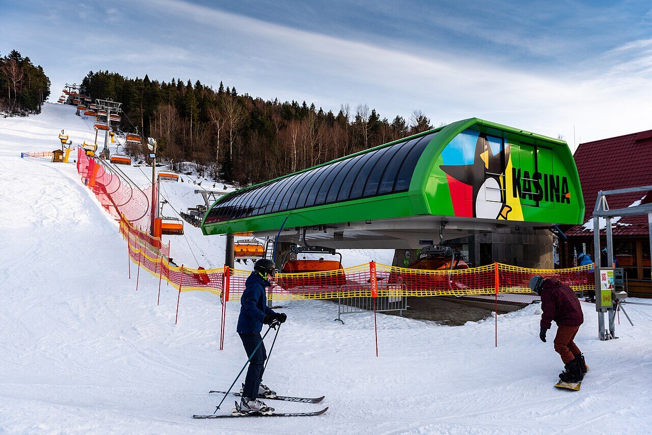 Stacja kolei krzesełkowej Kasina Ski — zielony budynek z logo, przed nim narciarz i snowboardzista na śniegu, w tle stok i las