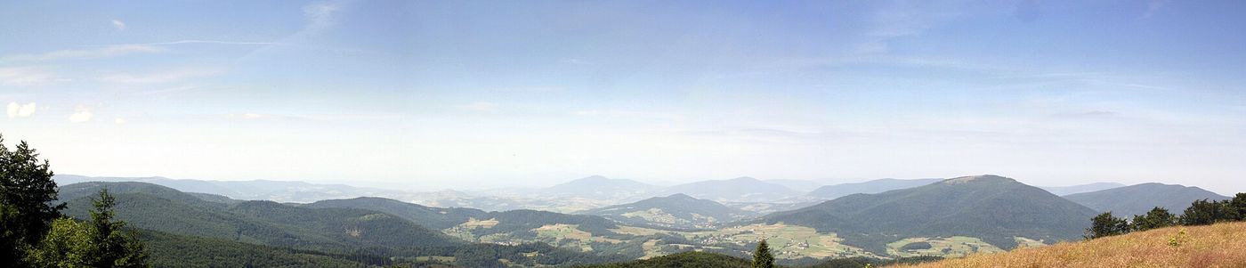Szeroka panorama ze szczytu Mogielicy - kilkadziesiąt kilometrów grzbietów Beskidu Wyspowego, doliny z wsiami i polami, na horyzoncie kolejne pasma w letnim zamgleniu. Po bokach kadru drzewa i trawa szczytowej polany. (ok. 4,6:1), typowy pas przez całą szerokość.