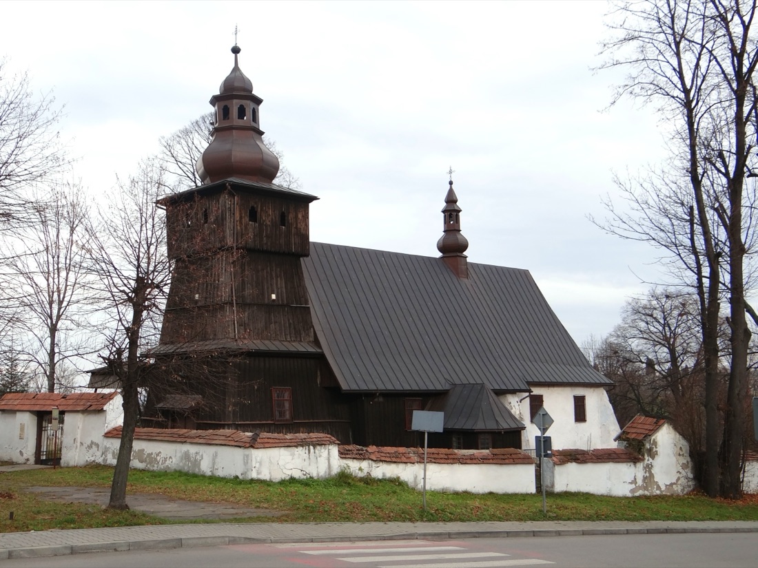 Widok od drogi, sprzed kamiennego muru — wieża z baniastym hełmem i długi dach nad nawą i murowanym prezbiterium.